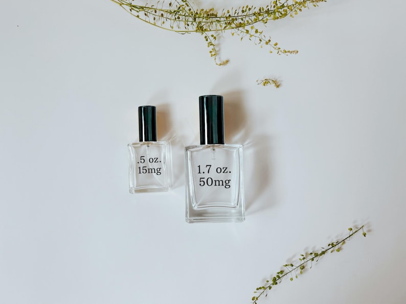 Peut inclure: Deux flacons de parfum en verre transparent avec des bouchons noirs, l'un &eacute;tiquet&eacute; ".5 oz. 15mg" et l'autre "1.7 oz. 50mg". Les flacons rectangulaires sont pos&eacute;s sur une surface blanche, avec des brins verts en arri&egrave;re-plan.