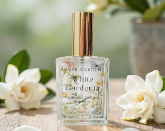 May include: A clear glass perfume bottle with a gold cap, featuring a label that reads Ember Gardens White Gardenia Handmade Perfume .5 oz | 15 mL. The label is decorated with a floral and butterfly print, and the bottle contains clear liquid.