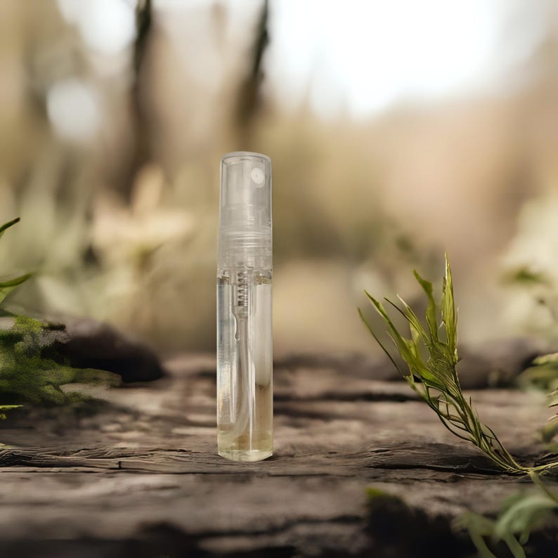 Peut inclure: Un flacon &eacute;chantillon de parfum en verre transparent avec un vaporisateur. Le flacon est rempli d'un liquide ambr&eacute; clair. L'arri&egrave;re-plan est flou, avec de la verdure et une surface en bois.