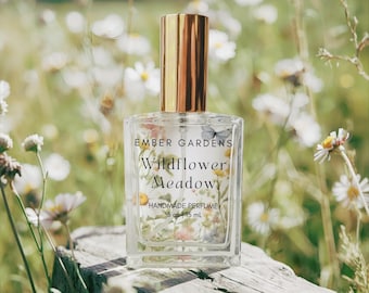 May include: A clear glass perfume bottle with a gold cap, labeled "Ember Gardens Wildflower Meadow Handmade Perfume." The bottle contains floral elements and is set against a blurred background of wildflowers. The bottle is 5 oz / 15 ml.