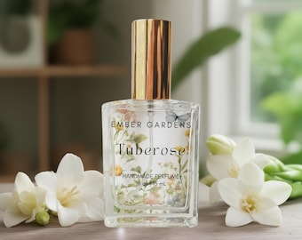 May include: A clear glass perfume bottle with a gold cap, labeled "Tuberose" and "Handmade Perfume." The bottle contains floral imagery. White flowers are arranged around the bottle. The bottle is 15 ml / .5 oz.