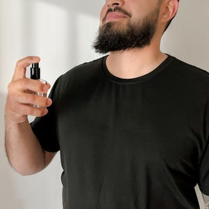 Peut inclure: Un flacon de parfum en verre transparent avec un vaporisateur noir est tenu. La personne porte un t-shirt noir. L'arri&egrave;re-plan est un mur blanc uni.