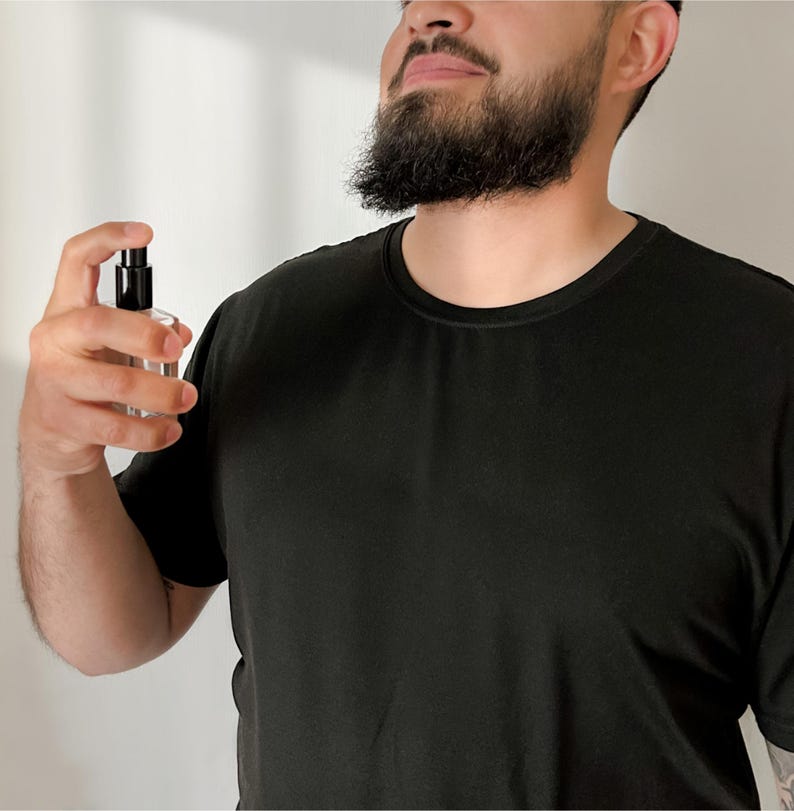 Peut inclure: Un flacon de parfum en verre transparent avec un vaporisateur noir est tenu. La personne porte un t-shirt noir. L'arri&egrave;re-plan est un mur blanc uni.