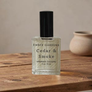 Peut inclure: Un flacon en verre transparent de cologne artisanale "Cedar & Smoke" avec un bouchon noir. L'&eacute;tiquette indique "Ember Gardens" et le volume : .5 oz | 15 ml. Le flacon est pos&eacute; sur une surface en bois.