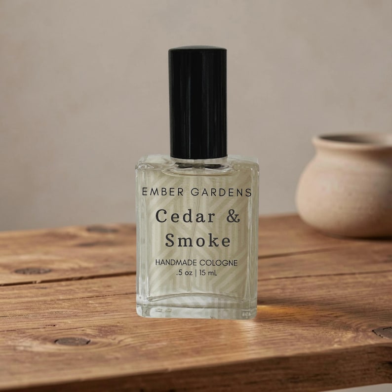 Peut inclure: Un flacon en verre transparent de cologne artisanale "Cedar & Smoke" avec un bouchon noir. L'&eacute;tiquette indique "Ember Gardens" et le volume : .5 oz | 15 ml. Le flacon est pos&eacute; sur une surface en bois.