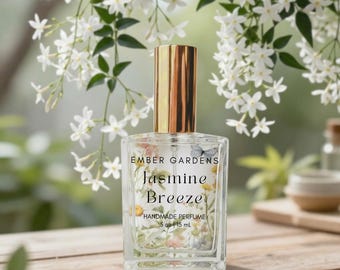 May include: A clear glass perfume bottle with a gold cap, labeled "Ember Gardens Jasmine Breeze Handmade Perfume." The bottle contains floral and butterfly designs. The background features white jasmine flowers and a wooden surface. The bottle contains 0.5 fl oz / 15 mL.