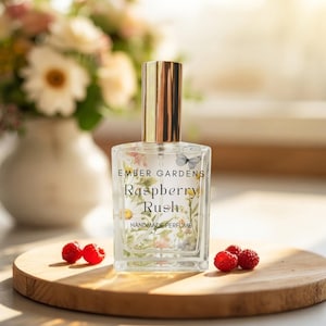 May include: A rectangular glass perfume bottle with a gold cap, featuring a label with the text Ember Gardens, Raspberry Rush, and Handmade Perfume, decorated with a floral and butterfly print, accompanied by four fresh red raspberries.