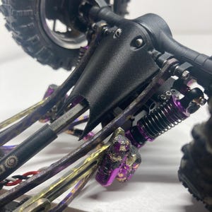 Puede incluir: Una placa protectora negra impresa en 3D montada en el eje trasero de un veh&iacute;culo teledirigido. El chasis cuenta con componentes de suspensi&oacute;n met&aacute;licos de color morado, amortiguadores negros y un eje de transmisi&oacute;n negro.