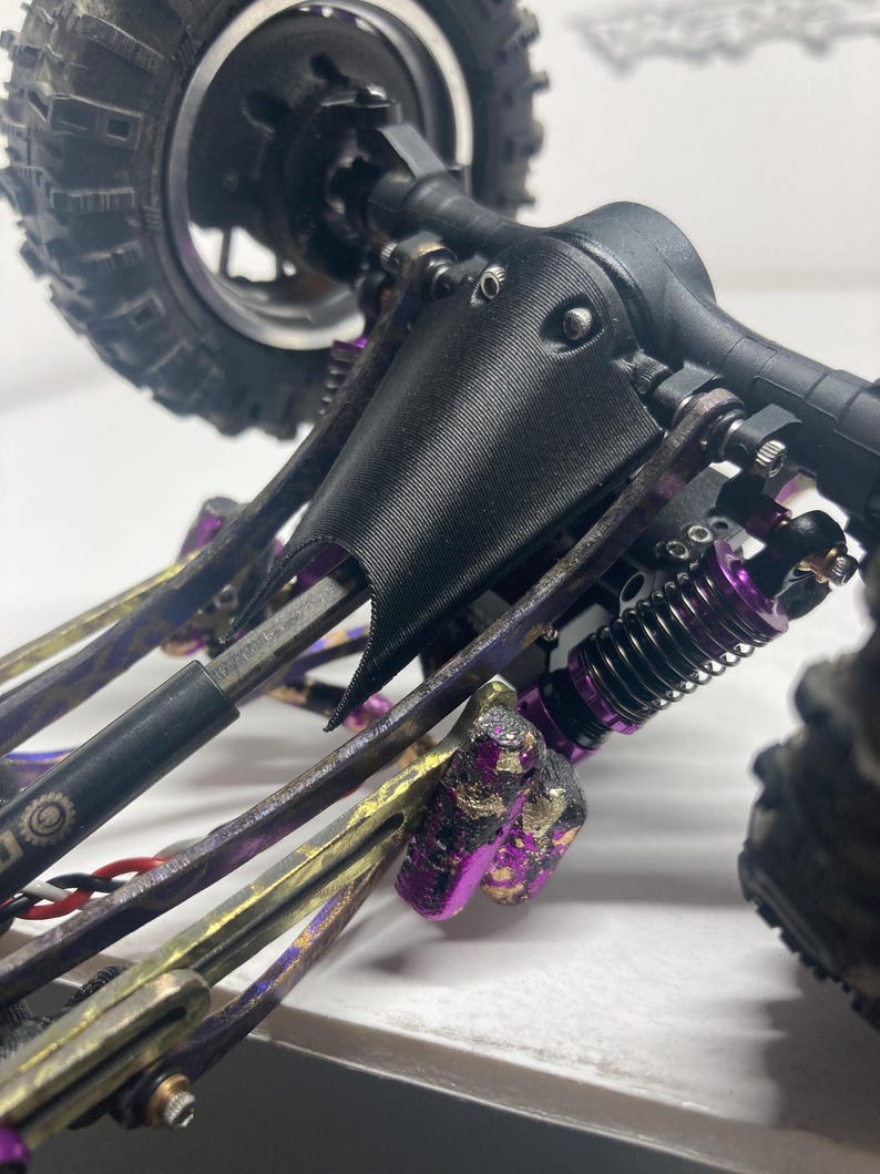Puede incluir: Una placa protectora negra impresa en 3D montada en el eje trasero de un veh&iacute;culo teledirigido. El chasis cuenta con componentes de suspensi&oacute;n met&aacute;licos de color morado, amortiguadores negros y un eje de transmisi&oacute;n negro.