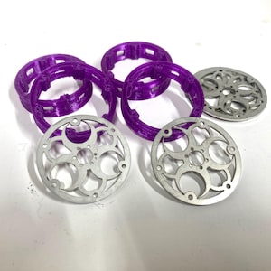 May include: A collection of circular components including four purple plastic rings with internal notched detailing and three silver-colored metal discs featuring a decorative cut-out pattern of interconnected circles and small circular holes.