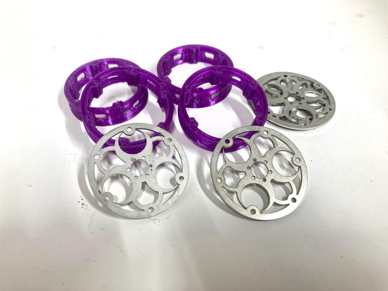 May include: A collection of circular components including four purple plastic rings with internal notched detailing and three silver-colored metal discs featuring a decorative cut-out pattern of interconnected circles and small circular holes.