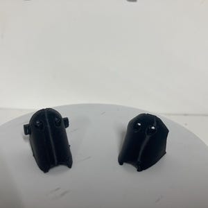 Puede incluir: Dos componentes de pl&aacute;stico negro impresos en 3D con una forma curva y c&oacute;nica, que presentan dos peque&ntilde;as hendiduras circulares cerca de la parte superior. Cada pieza tiene un acabado mate liso y una cresta vertical en el centro.