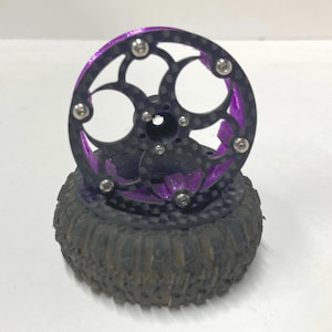 May include: A remote control vehicle wheel featuring a black carbon fiber rim with a circular cutout pattern, secured by silver metal screws. The inner structure is vibrant purple, and the tire is black rubber with a deep, rugged tread pattern.