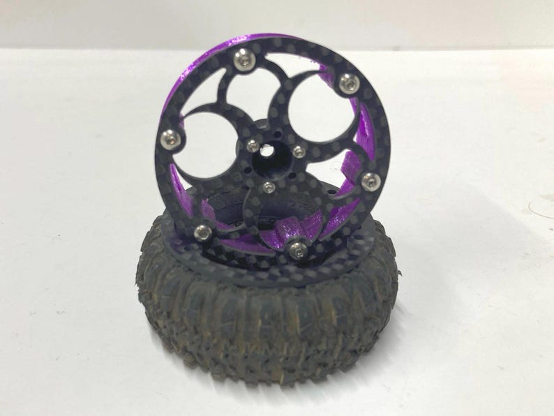 May include: A remote control vehicle wheel featuring a black carbon fiber rim with a circular cutout pattern, secured by silver metal screws. The inner structure is vibrant purple, and the tire is black rubber with a deep, rugged tread pattern.