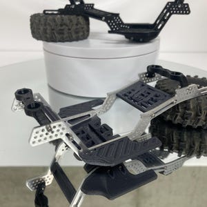 May include: Two black and silver model car chassis with tires. The chassis are made of metal and plastic, with intricate details. One chassis is on a white surface, and the other is on a reflective surface.