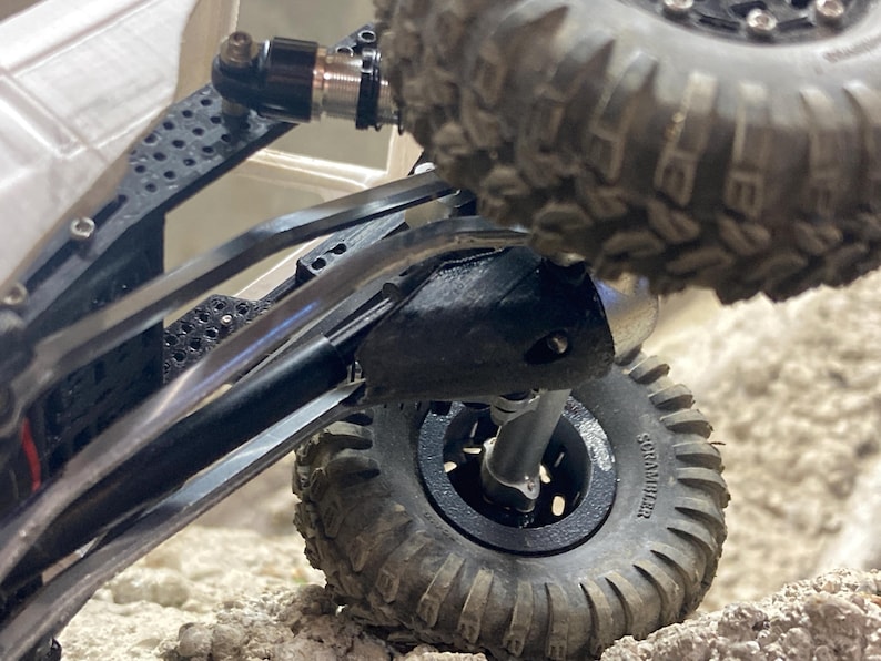 Puede incluir: Una vista de cerca del chasis de un veh&iacute;culo teledirigido tipo crawler con brazos de suspensi&oacute;n de fibra de carbono negra, un amortiguador met&aacute;lico y neum&aacute;ticos todoterreno negros con tacos profundos y la marca Scrambler.