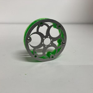May include: A circular object featuring a silver-colored metal faceplate with a decorative cutout pattern of interconnected circles and a central hole, attached to a bright green, textured, translucent base with visible screw fasteners.