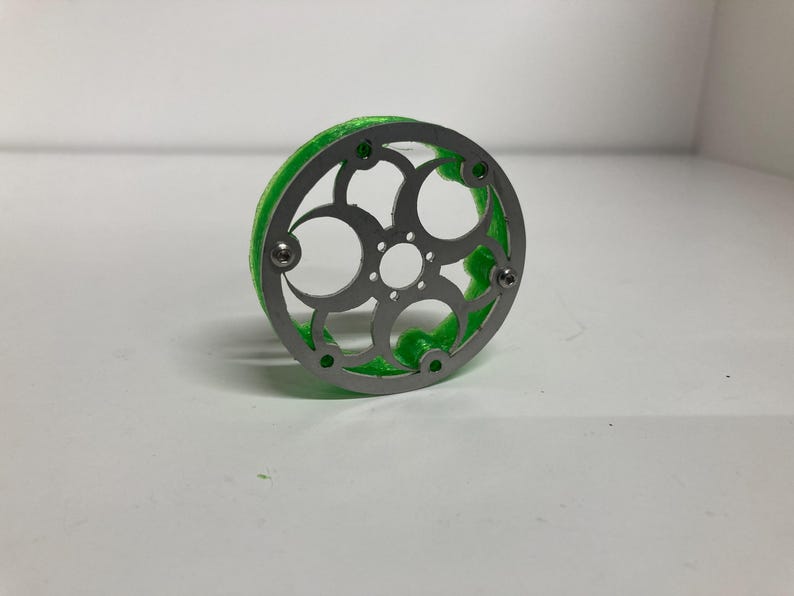 May include: A circular object featuring a silver-colored metal faceplate with a decorative cutout pattern of interconnected circles and a central hole, attached to a bright green, textured, translucent base with visible screw fasteners.