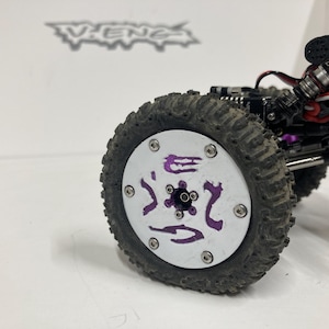 May include: A remote-controlled vehicle wheel featuring a white circular face with purple graphic accents and six silver screws. The tire is black with a textured tread pattern. The vehicle chassis includes black components, carbon fiber parts, and metal shocks.