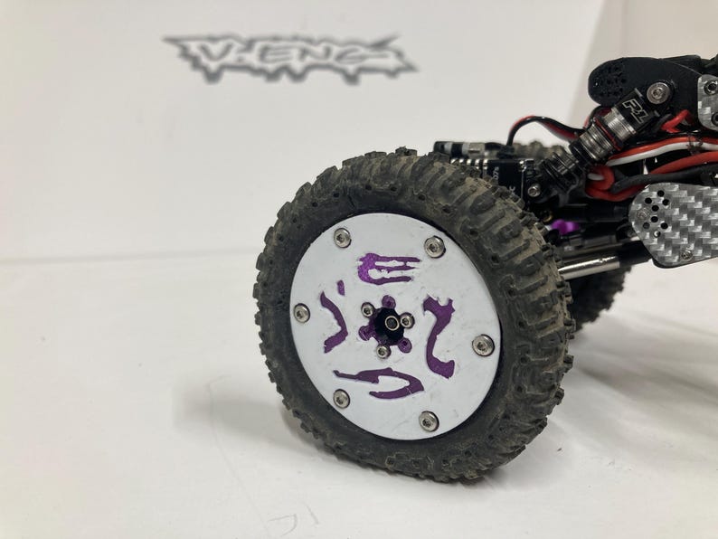 May include: A remote-controlled vehicle wheel featuring a white circular face with purple graphic accents and six silver screws. The tire is black with a textured tread pattern. The vehicle chassis includes black components, carbon fiber parts, and metal shocks.