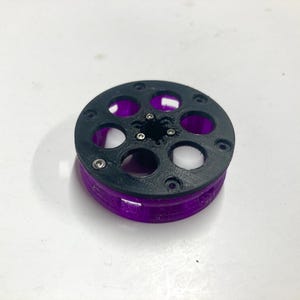 May include: A circular mechanical component featuring a black top plate with six circular cutouts and a central star-shaped opening. The base of the object is translucent purple with vertical supports, and small silver metal screws are visible.