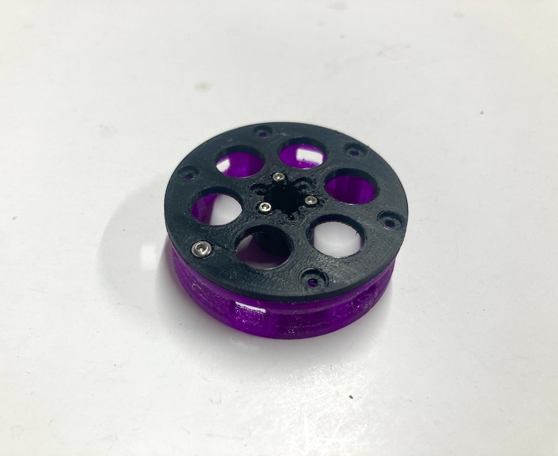 May include: A circular mechanical component featuring a black top plate with six circular cutouts and a central star-shaped opening. The base of the object is translucent purple with vertical supports, and small silver metal screws are visible.