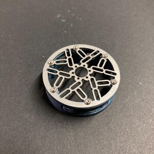May include: A circular component featuring a metallic silver top plate with a geometric cutout pattern of interconnected ovals and a central hole. The plate is secured by six small screws to a dark blue, textured cylindrical base.