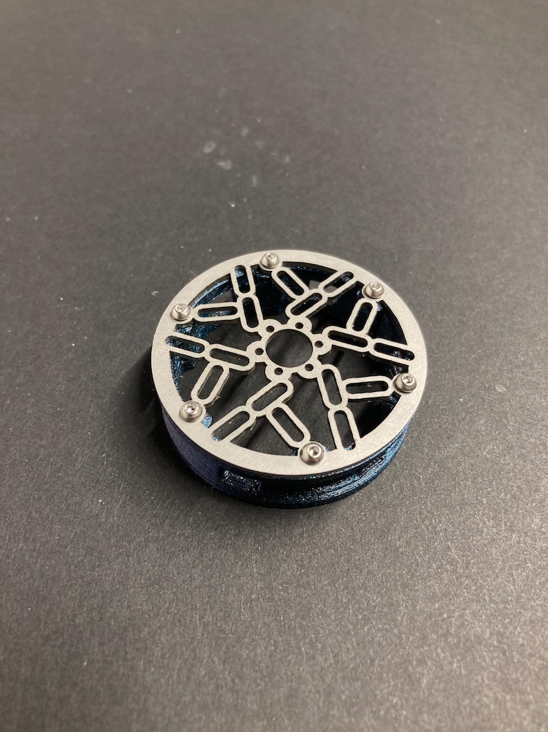 May include: A circular component featuring a metallic silver top plate with a geometric cutout pattern of interconnected ovals and a central hole. The plate is secured by six small screws to a dark blue, textured cylindrical base.