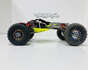 May include: A silver and black off-road RC car with yellow and red accents. The car has four wheels with black tires and silver rims. The car is on a white surface.