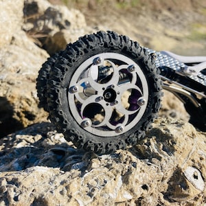 May include: A remote-controlled vehicle wheel featuring a silver metal rim with a circular cutout pattern, secured by six bolts. The tire is black with deep, rugged treads. The vehicle chassis is visible, showing carbon fiber and metal components.