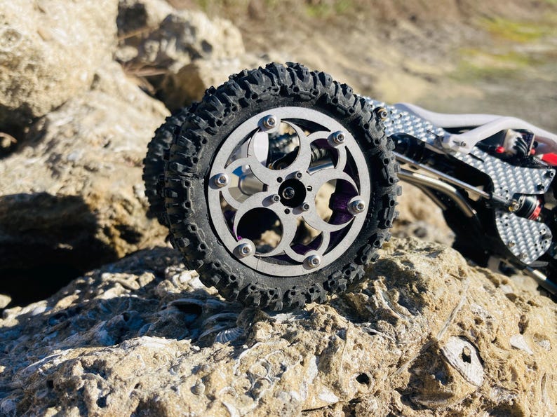 May include: A remote-controlled vehicle wheel featuring a silver metal rim with a circular cutout pattern, secured by six bolts. The tire is black with deep, rugged treads. The vehicle chassis is visible, showing carbon fiber and metal components.