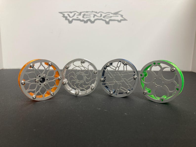 May include: Four circular fishing reels arranged in a row, featuring intricate metal frames with orange, silver, blue, and green accents. Each reel has a central hub and a complex, open-work spoke design secured with small silver screws.