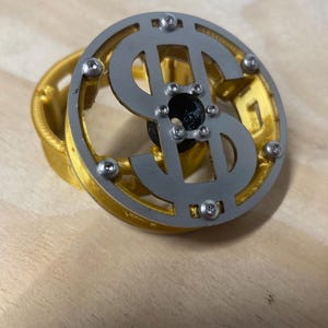 May include: A circular object featuring a silver-colored metal dollar sign emblem attached with small screws to a gold-colored plastic frame. The center of the dollar sign has a black circular component, and the entire assembly has a layered design.