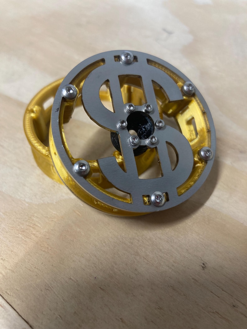 May include: A circular object featuring a silver-colored metal dollar sign emblem attached with small screws to a gold-colored plastic frame. The center of the dollar sign has a black circular component, and the entire assembly has a layered design.