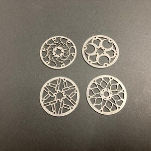 May include: Four circular, flat metal components featuring intricate, laser-cut geometric patterns. Each piece has a central hole surrounded by smaller perforations, with unique openwork designs radiating outward to the outer circular edge.