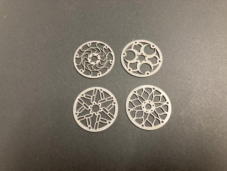 May include: Four circular, flat metal components featuring intricate, laser-cut geometric patterns. Each piece has a central hole surrounded by smaller perforations, with unique openwork designs radiating outward to the outer circular edge.