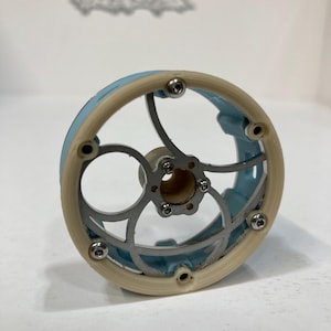 May include: A circular mechanical component featuring a light blue outer rim and a cream-colored frame. The center consists of a silver-toned metal structure with curved spokes and a central hub, secured by small silver screws around the perimeter.