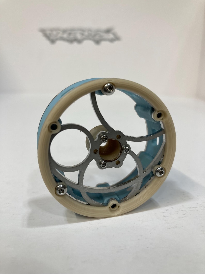 May include: A circular mechanical component featuring a light blue outer rim and a cream-colored frame. The center consists of a silver-toned metal structure with curved spokes and a central hub, secured by small silver screws around the perimeter.
