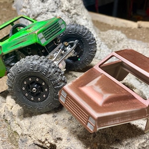 May include: A green and brown model truck body with a detailed interior and a weathered finish. The truck is missing its wheels and is sitting on a rock.