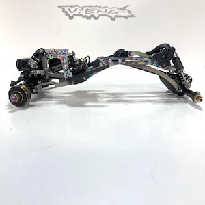 May include: A detailed view of a model car chassis, featuring a complex arrangement of metal components. The chassis has a black and iridescent finish, with visible shocks, springs, and other mechanical parts. The V-ENGS logo is visible in the background.
