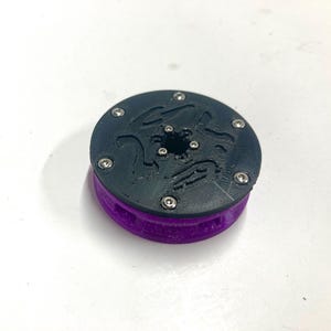 May include: A circular mechanical component featuring a black top plate with a textured surface and six small silver metal screws arranged in a ring. The base of the object is a vibrant purple color with a ribbed side profile and a central opening.
