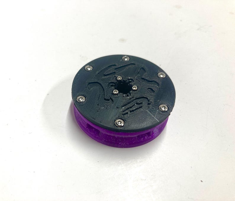 May include: A circular mechanical component featuring a black top plate with a textured surface and six small silver metal screws arranged in a ring. The base of the object is a vibrant purple color with a ribbed side profile and a central opening.