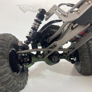 Pode incluir: Um chassis de veículo telecomandado com uma caixa de eixo verde, pneus todo-o-terreno pretos com rasto profundo e um sistema de suspensão em metal prateado com amortecedores de mola. A estrutura inclui parafusos visíveis e um motor.