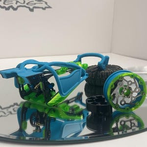 May include: A remote-controlled vehicle chassis featuring a bright blue frame and vibrant green internal components. The assembly includes a detached wheel with a green rim and a black tire, alongside a separate black circular hub component.