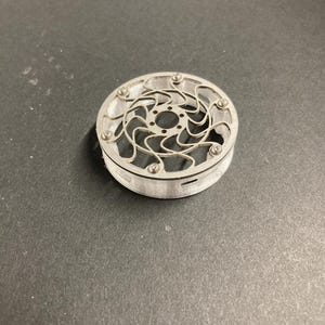 May include: A circular, silver-colored metal component featuring a complex, swirling, open-work pattern on its top surface. The piece is secured with small, evenly spaced screws along the outer rim and has a clear, cylindrical base.