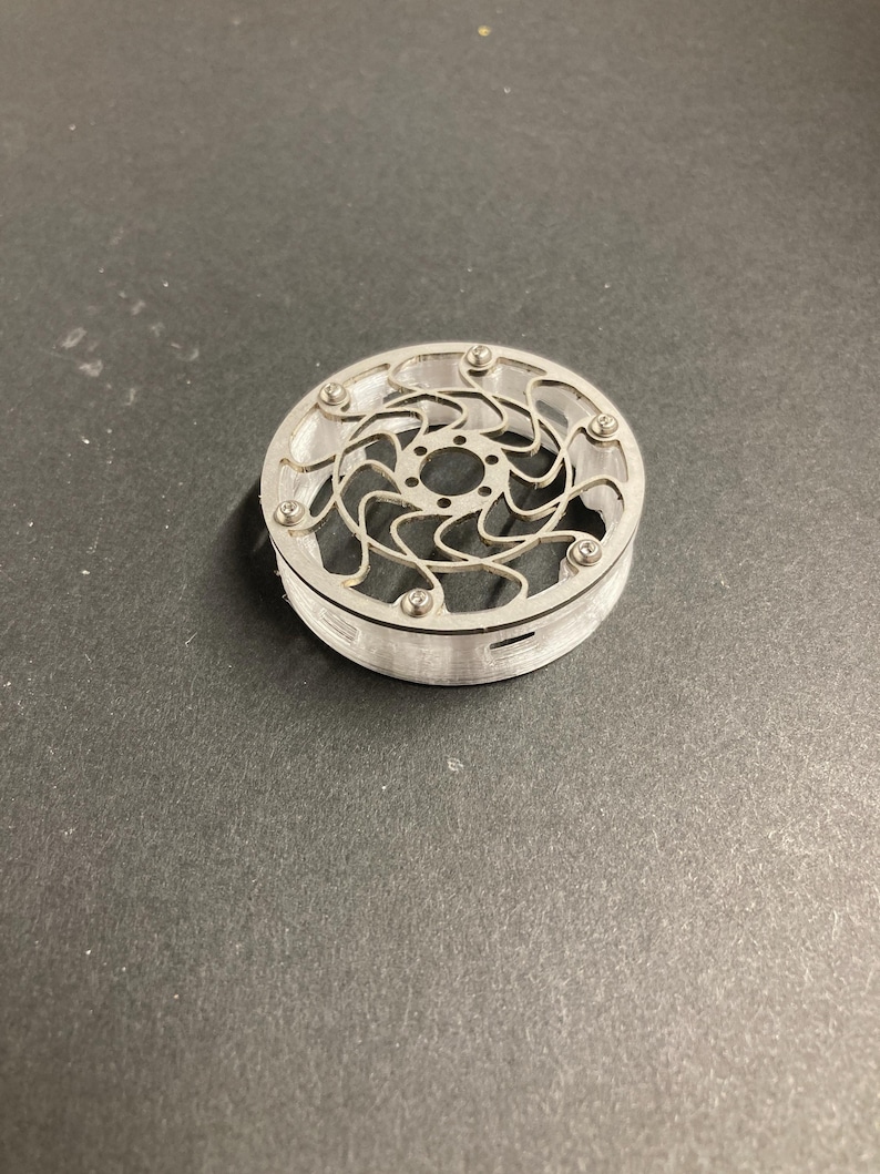 May include: A circular, silver-colored metal component featuring a complex, swirling, open-work pattern on its top surface. The piece is secured with small, evenly spaced screws along the outer rim and has a clear, cylindrical base.
