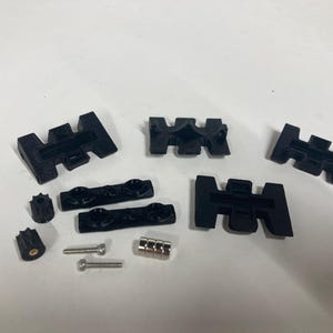 Puede incluir: Un surtido de componentes negros impresos en 3D, que incluyen piezas en forma de H, elementos cilíndricos y tornillos pequeños. Los artículos están dispuestos sobre una superficie blanca, lo que sugiere un conjunto de elementos mecánicos o estructurales.