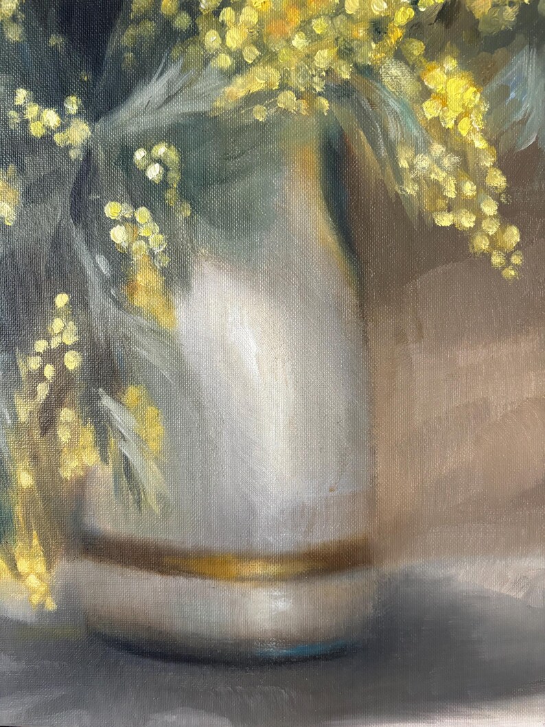 May include: A white ceramic vase with a gold band around the rim, filled with yellow mimosa flowers.