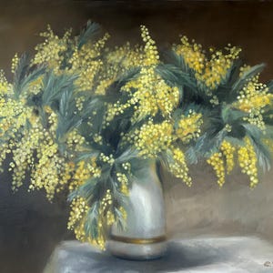 May include: A bouquet of yellow mimosa flowers in a white vase with a gold trim. The flowers are arranged in a loose, natural style and are set against a dark brown background.