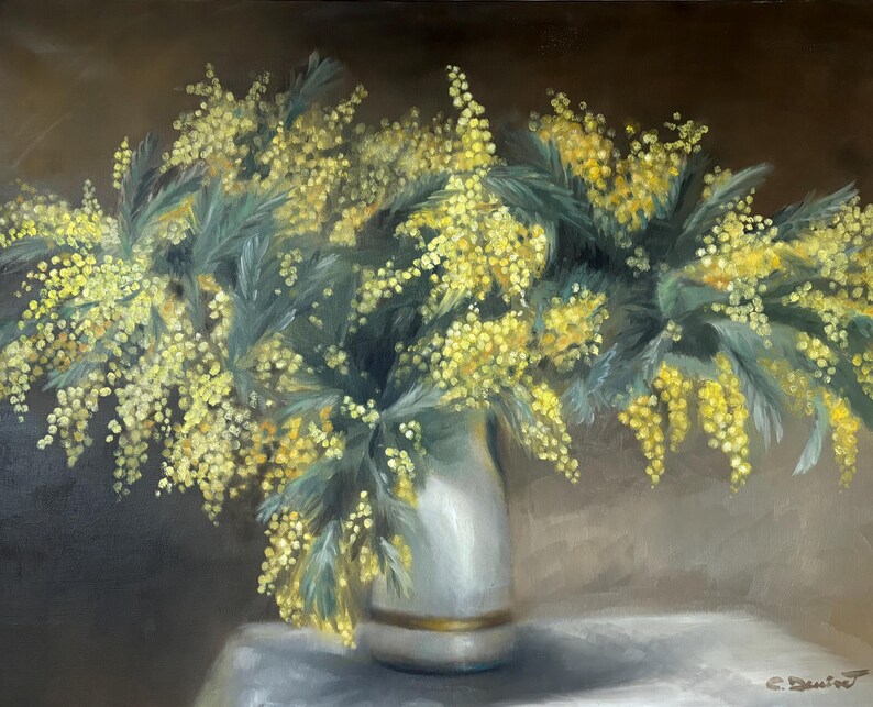May include: A bouquet of yellow mimosa flowers in a white vase with a gold trim. The flowers are arranged in a loose, natural style and are set against a dark brown background.